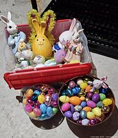 Red plastic container with assorted Easter bunny figurines, painted eggs, and decorative items inside. Two metal containers with colorful plastic eggs placed in front.