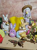 Overview of assorted Easter decorations including bunny figurines, yellow vase, purple bunny face decoration, butterfly ornament, and artificial flowers.