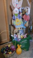 Photo showing four garden stake bunnies with white fabric ears and paint faces in shirts and straw hands and feet, with an Easter sign and ceramic vase.