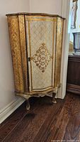 Front view of corner cabinet showing white painted body with gold gilt patterns, single door and cabriole legs.