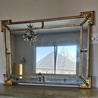 Large rectangular wall mirror with gold decorative frame placed on a surface against a wall. The frame features ornate corner decorations and beveled mirrored sections around the perimeter, reflecting a window and chandelier.
