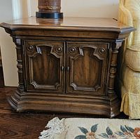 Solid wood end table with detailed paneled cupboard doors and decorative turned columns beside upholstered yellow couch. Wood has rich medium tone and beveled edge tabletop.