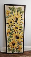 Full view of the framed sunflower needlepoint art leaning against a white wall on a floor baseboard.
