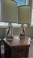 Two square-base table lamps with painted wild bird and greenery designs, topped with green fabric shades, placed on a wooden side table by a window.