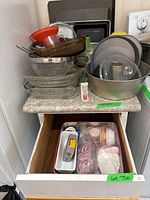 Overall assortment of metal and glass baking pans and trays stacked on countertop with drawer beneath showing muffin sleeves and kitchen jars inside.