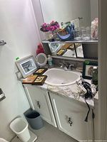 Photo of bathroom sink area with white cabinetry, marble countertop, three curling irons with cords, framed wall art, small trash cans, and a scale visible.