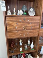 Shelf displaying multiple ceramic bells, figurines, a glass flower, and a battery powered clock face with wood grain design and metal numerals and hands. Small to medium size items.