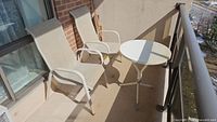 Two beige metal framed chairs with mesh fabric seating and backs, placed side by side on a balcony next to a round metal table with taped top and tripod base. Visible rust on all metal parts and peeling paint on table.