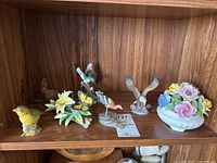 Seven figurines displayed on wooden shelf, showing variety including floral ceramics, birds, and carved wood bird