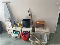 Wide shot showing two ironing boards, T-fal iron, plastic clothes baskets, cleaning liquids, white plastic tables, upholstered hamper, small wood stool, mop and broom sticks.