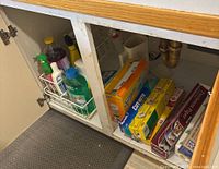 Under-sink wire rack with various cleaning bottles and boxed food wraps including foil, wax paper, parchment paper, and plastic wrap laid flat in cabinet.