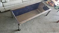Full view of the metal framed coffee table with mirrored glass top on carpet with visible chip in the glass corner and round metal legs.