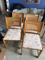 Four wooden dining chairs grouped together indoors on carpeted floor, showing the cane backs and floral upholstered seats.
