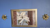 Frontal view of the framed floral art print with two gold-tone metal flowers mounted on each side against a blue wall.