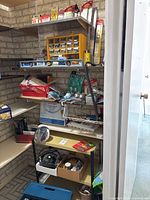 A storage corner with a small 24-inch metal shelving unit holding boxes, tools, cords, and other household items against a concrete block wall.
