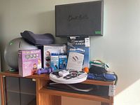 Photo showing full lot with massager, humidifier box, 3lb weights, exercise ball, wrist braces, back support, and other wellness items on wooden shelf and floor.