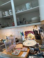 Kitchen cabinet with assorted glassware, cups, mugs, and dishes above a counter with canisters, trays, cutting board and a large floral plate.
