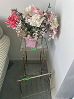 Full view showing metal plant stand with faux floral arrangement in ceramic pot on top shelf and metal magazine rack in front on floor next to plant stand