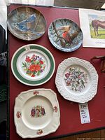 Six collector plates displayed on red surface: two bird plates, commemorative plates with royal imagery, floral prints, and embossed design.