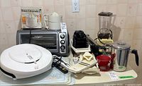 Photo showing Braun Citromatic 2 citrus juicer in box, Osterizer blender with plastic jug, Hamilton Beach toaster oven, Lean Mean Fat Reducing Grilling Machine, mandolin slicers, coffee percolator, fork thermometer, and cutting boards on kitchen counter.