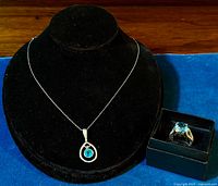 Photo showing the pendant necklace on a black display bust and the ring inside a small black box side by side on a blue cloth.