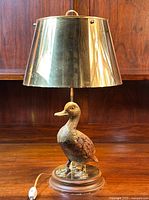 Full view of vintage table lamp showing brass shade, wooden duck figurine, and brass base on wooden surface.