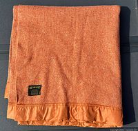 Folded burnt orange wool blend blanket with satin edging and black label showing Kenwood brand.