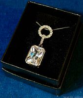 Pendant necklace displayed flat in a black box with black velvet lining: showing large rectangular clear cubic zirconium gemstone below a circle of smaller zirconium stones on a silver setting and chain.