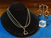 Front view of the multi-strand sterling silver necklace with floating heart pendant on a black display stand, alongside the black enamel and silver drop earrings on a clear stand.
