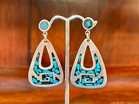 Front view of turquoise inlaid triangular sterling silver earrings hanging on a display stand showing the detailed turquoise mosaic pattern inlaid into the brushed silver surface.
