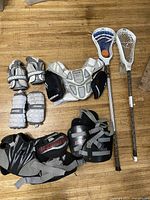 Overview of two lacrosse sticks and protective gear including gloves, pads, shoulder protector, and belt on wooden floor