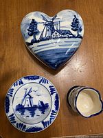 Top view of three Delft Blue ceramic pieces on wooden surface: heart-shaped trinket box, small plate, and mini vase or cup, all handpainted with blue Dutch windmill motifs.