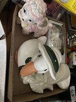 Photo showing four ceramic pieces including a pink poodle figurine and a white duck figurine with a green hat, along with two other white ceramic pieces.