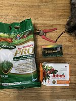 Photo showing all four items in the lot: Scotts lawn food bag, Jobe's fertilizer spikes, SunMate Plus sprinkler nozzle, and red pruners on a wooden surface with some pet paw visible.