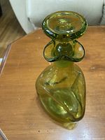 Green glass decanter shown from left angled front view on wooden table with cream couch in background.