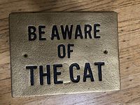 Front view of rectangular gold-tone cast iron sign with bold black lettering stating 'BE AWARE OF THE CAT' and wall mounting holes.