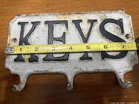 Front view of cast iron key holder showing raised black 'KEYS' letters on white background with attached measuring tape indicating approximately 7 inches wide and three hooks for keys.