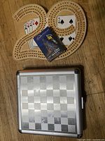 Wooden cribbage board with small pack of cards placed on top, next to a closed metal travel chess case with a checkered pattern.