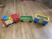 Full view of all three toy train pieces including the red locomotive, green flatbed car, and blue/yellow caboose on wood floor.