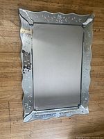 Front view of rectangular hanging wall mirror showing etched floral border and scalloped edge design on wood floor background.