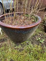 Photo of a large ceramic pot with plants and soil inside, showing glazed brown and blue patina and resting on grassy ground outside.