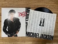 Michael Jackson Bad album vinyl record, outer cover, and lyrics insert sheet displayed on wooden surface.