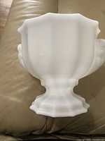Side view of white milk glass pedestal vase showing fluted body, two rounded handles, scalloped edges on base and rim.