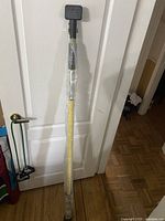 Full view of the Quick Support Rod wrapped in plastic, showing rubber top and cushioned handle, propped against a white door.