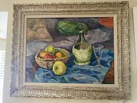Framed oil painting showing a still life with basket of fruit, bottle, and glass on blue cloth