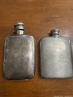Two pewter flasks side by side on a wooden surface, showing overall condition and size difference.