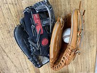 Two baseball gloves and one baseball on wooden floor, one black/red glove worn, one tan Wilson glove good condition, one baseball included.