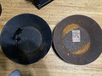 Two round gold pans placed on a wooden surface: one black, one rusted