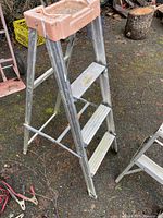 Full view showing the 4-foot aluminum step ladder with four ridged steps and an orange plastic top tray, standing outdoors on dirt ground.