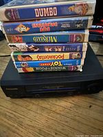 Stack of seven Walt Disney VHS movies on top of a black Panasonic VHS player showing front controls and cassette slot, placed on a wooden surface.